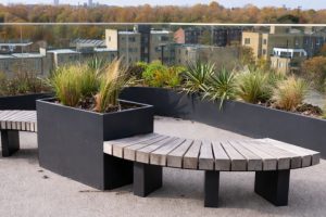 Anthracite aluminium planters installed on a rooftop terrace with integrated wooden bench seating and mixed planting overlooking city buildings