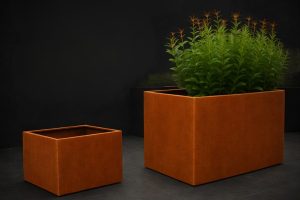 Two corten steel cube metal planters with warm rust finish placed on dark tiled floor, one planted with green ornamental plants and one empty