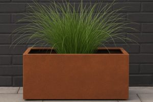 Corten steel rectangular trough planter with rust finish placed against dark brick wall, filled with tall ornamental grass
