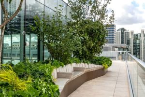 Curved metal planter with integrated bench seating and lush planting on a modern UK rooftop terrace with glass facade