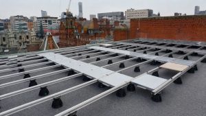 Metal planter roof terrace subframe system with aluminium rails, adjustable pedestal supports and paving panels being installed on a UK city rooftop.