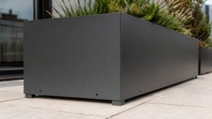 Close-up detail of a powder-coated anthracite aluminium metal planter on a UK rooftop terrace, showing smooth matte finish, drainage holes and raised feet above pale paving.